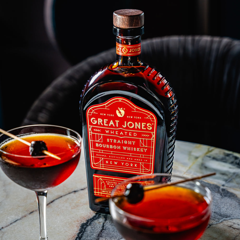 
                  
                    Bottle of Great Jones Wheated Straight Bourbon Whiskey on a marble table, alongside two Manhattan cocktails served in coupe glasses with cherry garnishes
                  
                