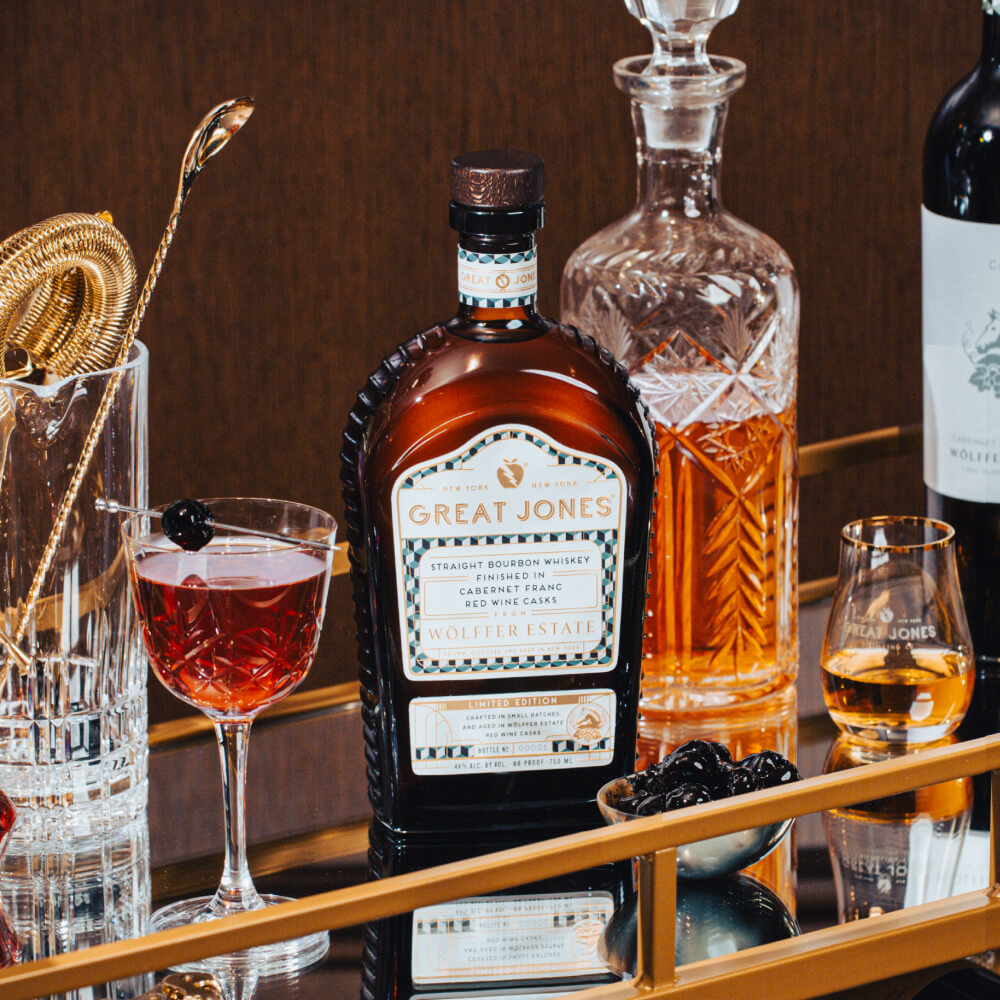 Bottle of Great Jones x Wölffer Estate Cask Finished Bourbon on a mirrored bar cart surface, surrounded by a Manhattan cocktail and barware