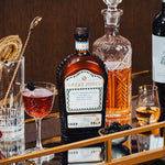 Bottle of Great Jones x Wölffer Estate Cask Finished Bourbon on a mirrored bar cart surface, surrounded by a Manhattan cocktail and barware